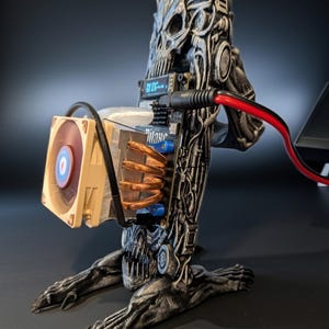 Peut inclure: Un boîtier d'ordinateur personnalisé en forme de crâne, avec des détails complexes et une finition métallique. Le boîtier intègre un ventilateur, une carte de circuit imprimé et d'autres composants informatiques. Un câble rouge et noir sort du boîtier.