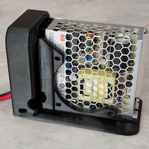 May include: A black 3D-printed plastic mounting bracket holding a silver metal power supply unit. The power supply features a hexagonal mesh cover, internal electronic components, and a label with the text MW LRS-350-24 visible on the side.