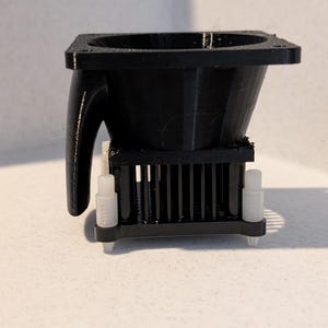 May include: A black plastic component with a square top and a curved handle. The component has a series of vertical bars and is supported by four white cylindrical posts. The object is likely a part of a larger mechanism.
