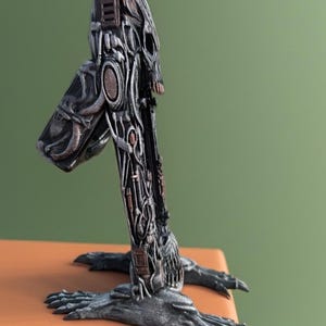 Könnte beinhalten: Eine detaillierte Metallskulptur mit einem mechanischen, futuristischen Design. Die Skulptur zeigt eine kopfartige Struktur mit komplizierten Details, einen langen Hals und klauenartige Füße. Die Farbpalette umfasst Grautöne, Silber und Bronze.