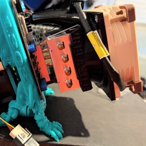 May include: A computer cooling component featuring a metallic heatsink with four copper heat pipes, a black fan shroud labelled VRBLASTER, and a tan fan. It is attached to a bright teal 3D-printed structure with the text 1303 visible on the side.
