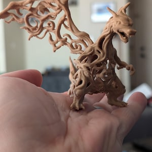 Op de afbeelding: Een lichtbruin 3D-geprint beeldje van een draakachtig wezen met grote, ingewikkelde, wervelende vleugelstructuren en een getextureerd, organisch oppervlak. Het wezen staat op twee poten met een open mond en puntige hoorns op de kop.