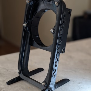 May include: A black, 3D-printed stand with a circular opening and the text "6xQe NerdCave" on the side. The stand has a base with two feet and is held together with silver screws.