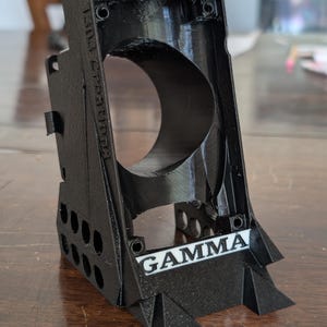 May include: Black 3D-printed object with the text "BITAXE" and "GAMMA" in white. The object has a large circular opening and several smaller holes. The words "1303 creations" are also visible.