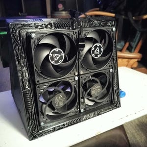 May include: A black, custom-built computer cooling component featuring four square Arctic brand fans arranged in a two-by-two grid. The housing has a textured, industrial-style frame with intricate, raised linear patterns surrounding the fans.