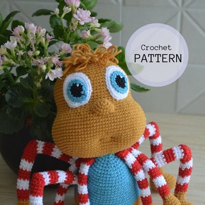 Può includere: Giocattolo amigurumi a forma di ragno lavorato a uncinetto con gambe a strisce rosso e bianco, camicia blu e corpo marrone. Il giocattolo ha grandi occhi blu con pupille nere. Schema all'uncinetto.