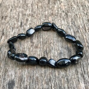 May include: A black beaded bracelet composed of oval-shaped stones. The bracelet is displayed on a weathered wooden surface, showcasing the dark, polished beads. The bracelet is a piece of jewelry.