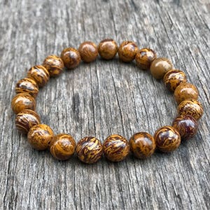 May include: A beaded bracelet featuring round, polished beads in shades of brown and gold with unique patterns. The bracelet is made of natural stone and is displayed on a weathered wooden surface. A stylish accessory.