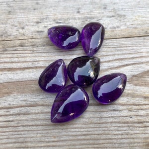May include: Six purple amethyst teardrop shaped stones on a wooden surface.