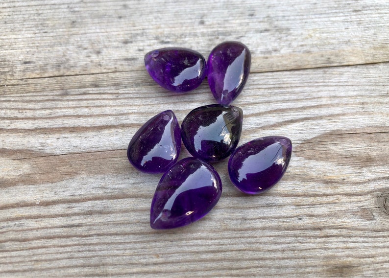 May include: Six purple amethyst teardrop shaped stones on a wooden surface.