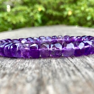 May include: A faceted amethyst bracelet. The bracelet is a deep purple color and is made of multiple, small, geometric-shaped beads strung together. The bracelet is resting on a weathered wooden surface, with a blurred green background.