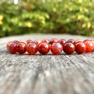 Puede incluir: Una pulsera de cuentas redondas y pulidas en tonos rojos y naranjas. La pulsera descansa sobre una superficie de madera envejecida, con un fondo verde borroso.