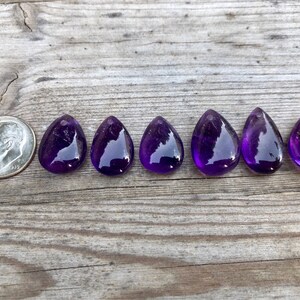 May include: Seven teardrop-shaped amethyst gemstones, each approximately the size of a US dime, arranged in a row on a wooden surface.