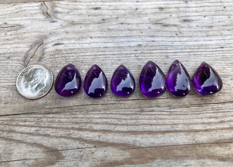 May include: Seven teardrop-shaped amethyst gemstones, each approximately the size of a US dime, arranged in a row on a wooden surface.