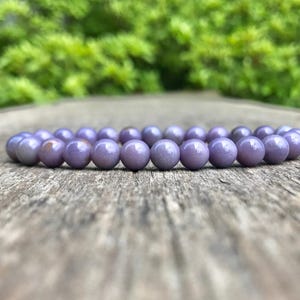 May include: A lavender beaded bracelet rests on a weathered wooden surface. The bracelet is composed of numerous round, smooth beads. The background features blurred green foliage, creating a natural aesthetic. The bracelet is a piece of jewelry.