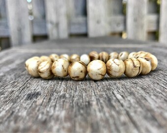 Könnte beinhalten: Ein Armband aus kugelförmigen, cremefarbenen und braunen Perlen. Das Armband liegt auf einer verwitterten Holzoberfläche. Die Perlen haben ein natürliches Aussehen, was auf ein handgefertigtes Produkt hindeutet. Der Umfang beträgt ca. 18 cm.