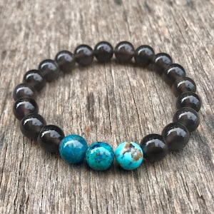May include: A beaded bracelet featuring dark gray, round beads and three turquoise-colored beads. The bracelet is displayed on a weathered, wooden surface, showcasing the contrast between the smooth beads and the textured background. This accessory is suitable for everyday wear.