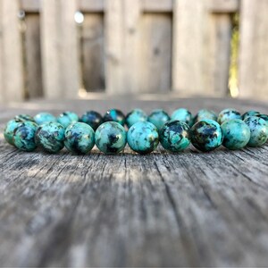 May include: A turquoise and black beaded bracelet. The round beads are a mix of light and dark turquoise with black marbling. The bracelet is displayed on a weathered wooden surface.