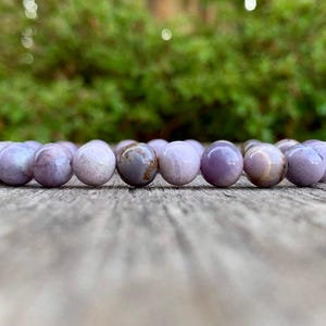 May include: A beaded bracelet featuring round, lavender-colored beads. The beads have a smooth, polished surface and are strung together on an elastic band. The bracelet is resting on a weathered wooden surface, with a blurred green background.