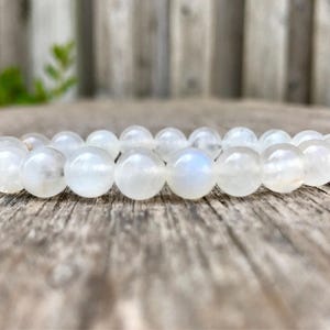 May include: A white moonstone bead bracelet. The bracelet is made of round, polished beads with a subtle iridescent sheen. The beads are strung together on an elastic band. The bracelet is resting on a weathered wooden surface.