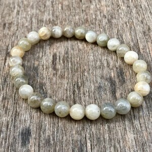 May include: A beaded bracelet composed of round, light green and white stones. The bracelet is strung together, forming a complete circle. The beads have a smooth, polished appearance. The bracelet is displayed on a wooden surface.