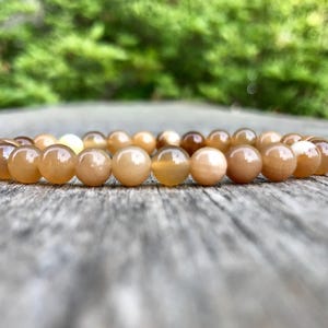 以下が含まれることがあります： ベージュ、茶色、クリーム色の丸く磨かれたビーズのブレスレット。ブレスレットは、風化した木製の表面に置かれ、背景にはぼやけた緑色の葉があります。