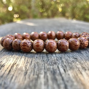 May include: A beaded bracelet featuring round, brown beads with a speckled pattern. The bracelet is displayed on a weathered wooden surface, with a blurred natural background.