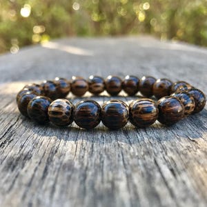 May include: A beaded bracelet with dark brown and tan wooden beads. The bracelet is made of round beads with a natural wood grain pattern. The bracelet is resting on a weathered wooden surface, with sunlight in the background.