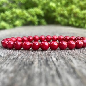 Puede incluir: Un brazalete de cuentas rojas, compuesto por numerosas cuentas redondas y brillantes, descansa sobre una superficie de madera envejecida. El fondo presenta un follaje verde borroso. Esta joya es adecuada para el uso diario. Las cuentas miden unos 0,8 cm de diámetro.
