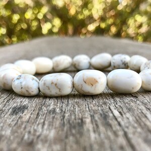 May include: A white stone bead bracelet with a natural, organic look. The smooth, oval-shaped beads have subtle variations in color and texture, creating a unique and elegant accessory. The bracelet is displayed on a weathered wooden surface.