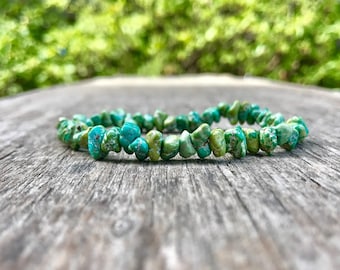 May include: A turquoise chip bead bracelet sits on a weathered wooden surface. The bracelet features irregular, light green and turquoise-coloured stones strung together. The background is a blurred view of green foliage.