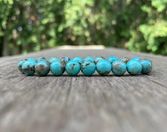 May include: A turquoise beaded bracelet with brown marbling. The round beads are strung together, resting on a weathered wooden surface. The background is blurred with green foliage.