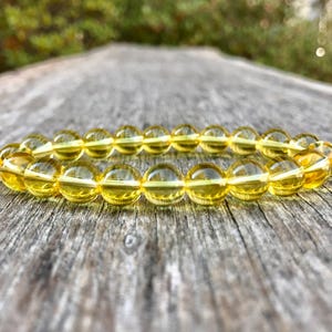 May include: A yellow beaded bracelet with translucent, spherical beads. The bracelet is resting on a weathered, gray wooden surface, with a blurred green background. The beads appear to be made of a clear, amber-colored material.