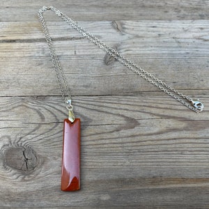 May include: A long, rectangular pendant made of brown stone hangs from a silver chain. The pendant has a smooth, polished surface and a slightly rounded edge.