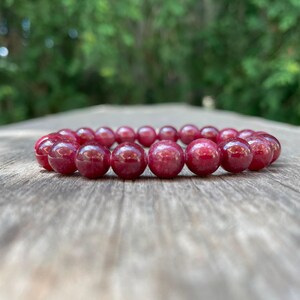May include: A beaded bracelet featuring large, round, polished beads in a deep red hue. The bracelet is resting on a weathered wooden surface, with a blurred green background of foliage.