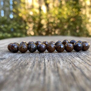 May include: A beaded bracelet featuring dark brown, faceted beads. The beads have a textured surface and are arranged in a circular pattern. The bracelet is displayed on a weathered wooden surface, with a blurred green background.