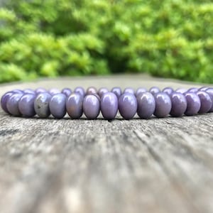 May include: A lavender beaded bracelet resting on a weathered wooden surface. The bracelet is composed of oval-shaped beads, each approximately 0.5 inches in length. The background features blurred green foliage, creating a natural aesthetic. This jewelry item is suitable for everyday wear.
