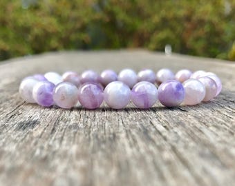 Puede incluir: Un brazalete de cuentas redondas y pulidas en tonos lavanda y blanco. El brazalete descansa sobre una superficie de madera envejecida, con un fondo verde borroso.