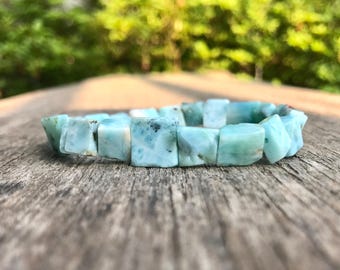 Puede incluir: Un brazalete de larimar azul claro con cuentas cuadradas. El brazalete está hecho de piedra natural y descansa sobre una superficie de madera envejecida. El fondo está borroso, mostrando follaje verde.