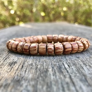 May include: A wooden bracelet with a natural, earthy aesthetic. The bracelet is composed of numerous small, cylindrical beads, each displaying a pattern of light brown and darker brown stripes. The bracelet rests on a weathered wooden surface, with a blurred green background.