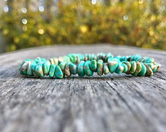 May include: A turquoise chip bead bracelet lies on a weathered wooden surface. The bracelet is made of irregular, light blue-green beads with brown and tan accents. The background is blurred, with hints of green and yellow foliage.