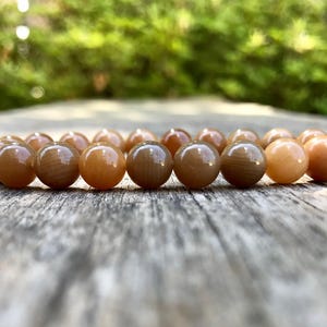 以下が含まれることがあります： 茶色とベージュの色合いの、丸く磨かれたビーズで構成されたブレスレット。 ブレスレットは、風化した木製の表面に置かれており、背景はぼやけた緑色です。