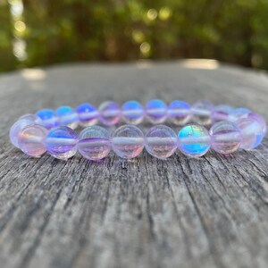 Puede incluir: Una pulsera de cuentas redondas e iridiscentes. Las cuentas cambian de color, mostrando tonos de azul, morado y rosa. La pulsera descansa sobre una superficie de madera envejecida, con un fondo verde borroso.