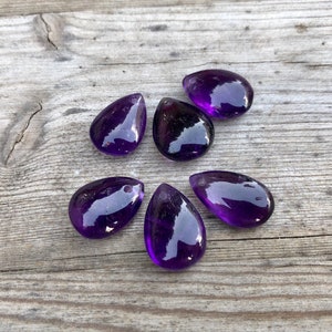May include: Six purple amethyst teardrop shaped gemstone pendants on a wooden surface.
