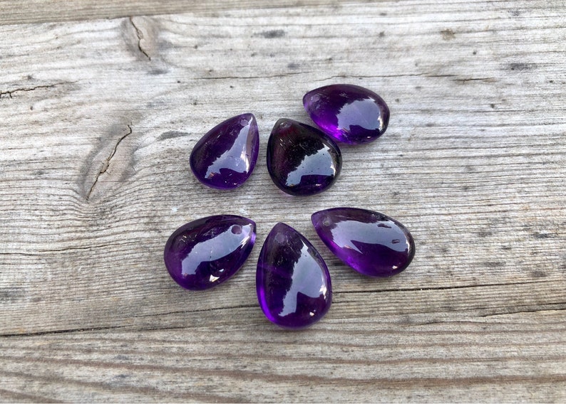 May include: Six purple amethyst teardrop shaped gemstone pendants on a wooden surface.
