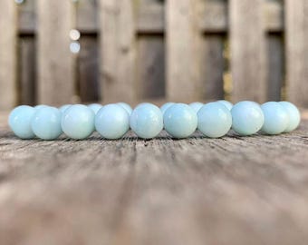 May include: A light blue beaded bracelet resting on a wooden surface. The bracelet is composed of round, smooth beads. The background features a wooden fence, with the focus on the bracelet's color and texture.
