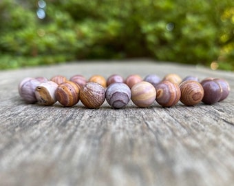 May include: A beaded bracelet featuring a variety of earth-toned beads. The beads are primarily shades of brown, tan, and purple, with a mix of textures and patterns. The bracelet is resting on a weathered wooden surface.