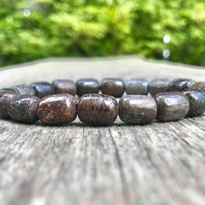 May include: A dark brown beaded bracelet with square-shaped beads. The bracelet is made of a natural stone material, with variations in color and texture. The beads are strung together to form a circular shape. The bracelet is resting on a wooden surface.
