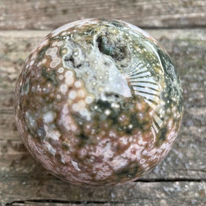 May include: A polished, green, pink, and white ocean jasper sphere. The sphere has a swirling pattern of colours and textures.