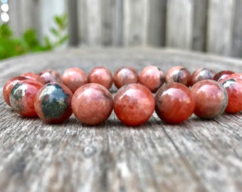 May include: A beaded bracelet featuring round, polished beads in shades of red, pink, and brown. Some beads have green and black inclusions. The bracelet is displayed on a weathered wooden surface, showcasing the natural stone beads.
